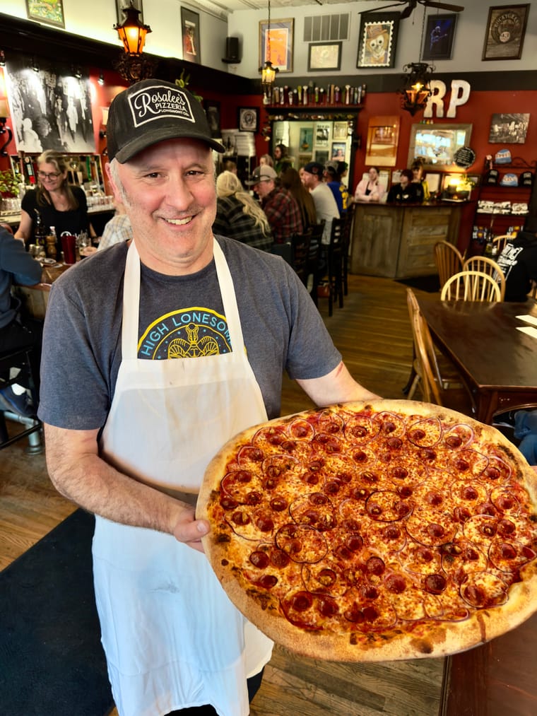 Owner of Rosalee's Pizzeria holding a large pepperoni pizza inside the restaurant